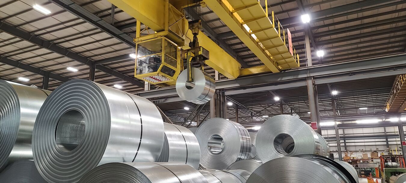 Stacks of galvanized coil on the facility floor with a coil being lifted by a yellow crane.