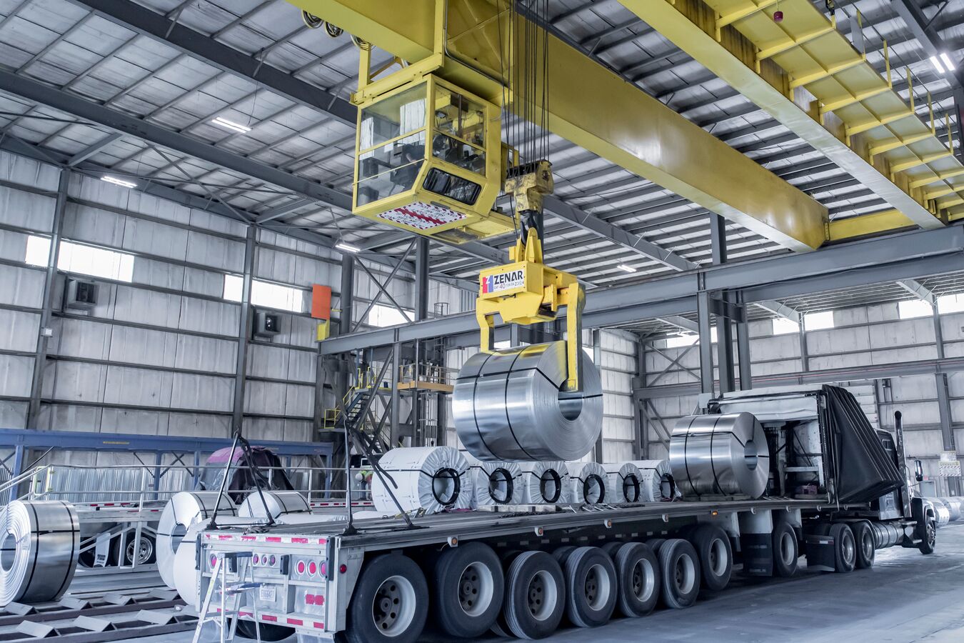 A master coil of galvanized steel being lowered from a yellow crane onto a flatbed truck for delivery.