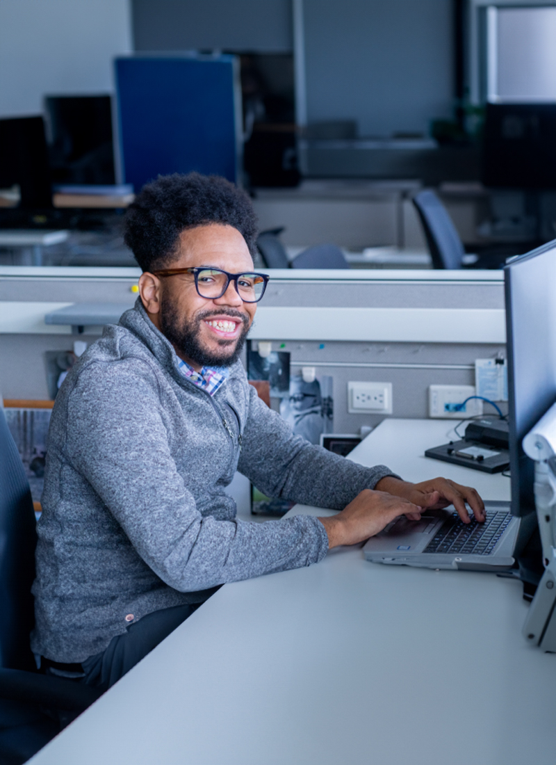A closeup of a male employee smiling, sitting at a desk and working on a laptop.