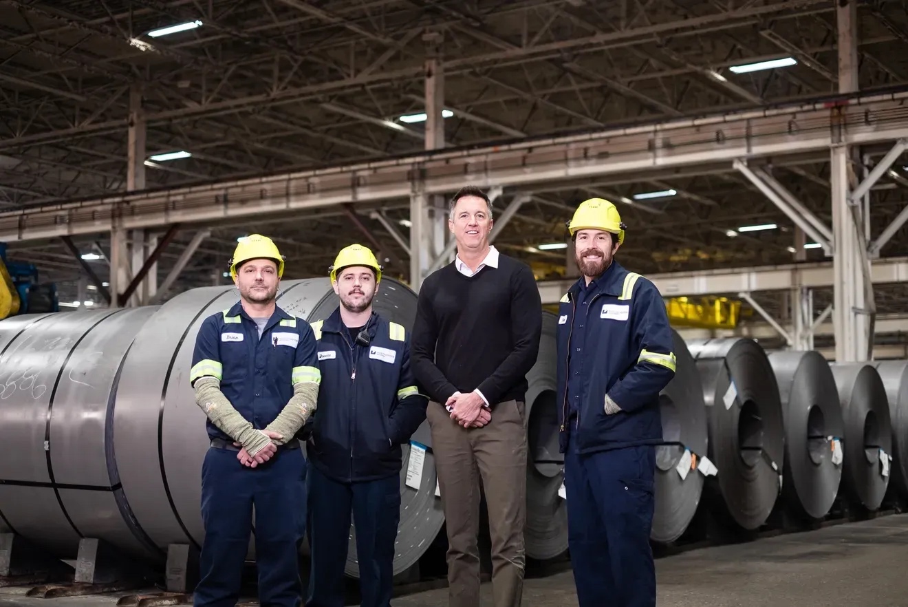 A group of Worthington Steel employees in front of steel coils smiling at the camera with PPE on.