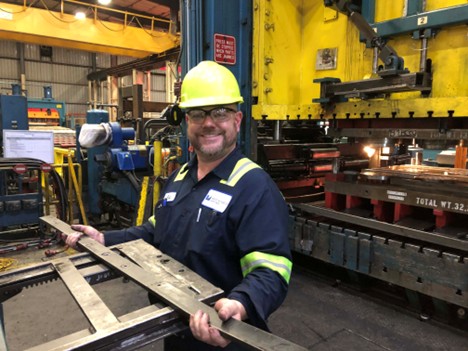 An employee on the manufacturing floor, wearing safety gear, and holding a steel part.